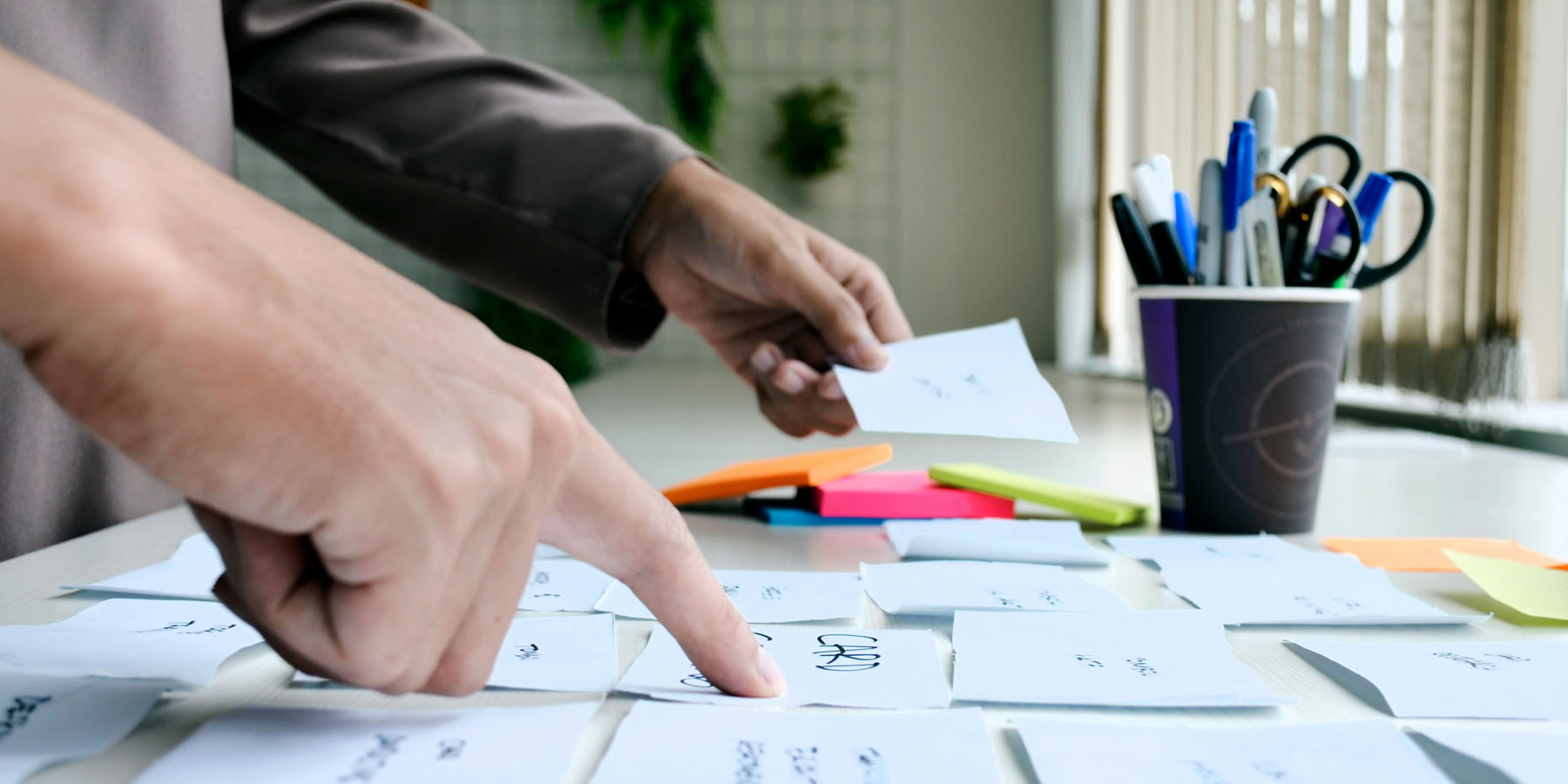 Hands sorting through index cards on a table, with a cup holding pens and pencils in the background.