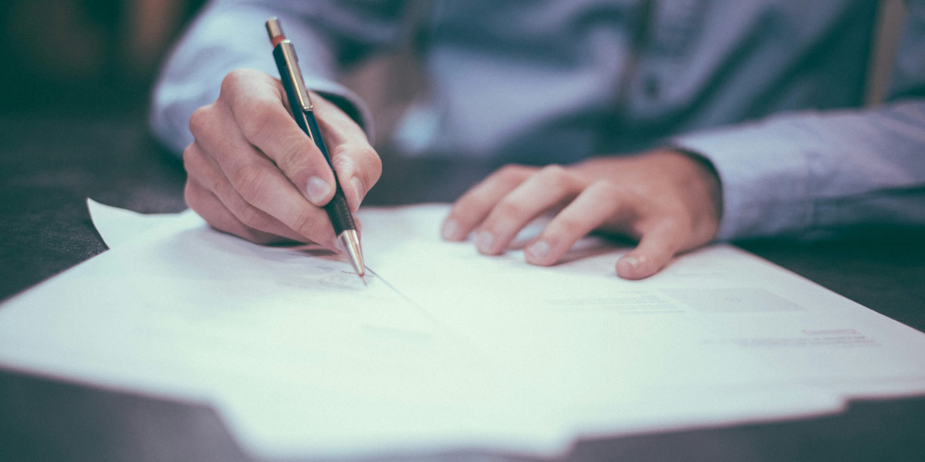 A person in a blue shirt writing on papers with a pen on a desk.
